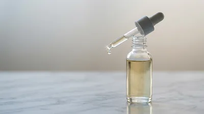 A single unbranded clear glass dropper bottle of pale golden serum on a polished marble surface, a droplet suspended at the dropper tip in clean cool light against a soft grey and champagne background
