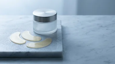 Two unbranded crescent-shaped translucent gel eye patches resting on a chilled pale grey marble slab beside a plain frosted glass jar with a brushed steel lid, in cool morning light