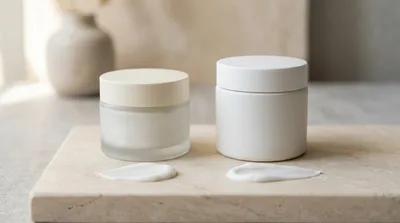 Two unbranded cream jars side by side on a pale travertine slab, one squat frosted glass jar with a cream lid and one taller matte white ceramic jar, each with a small swatch of white cream beside it in soft daylight